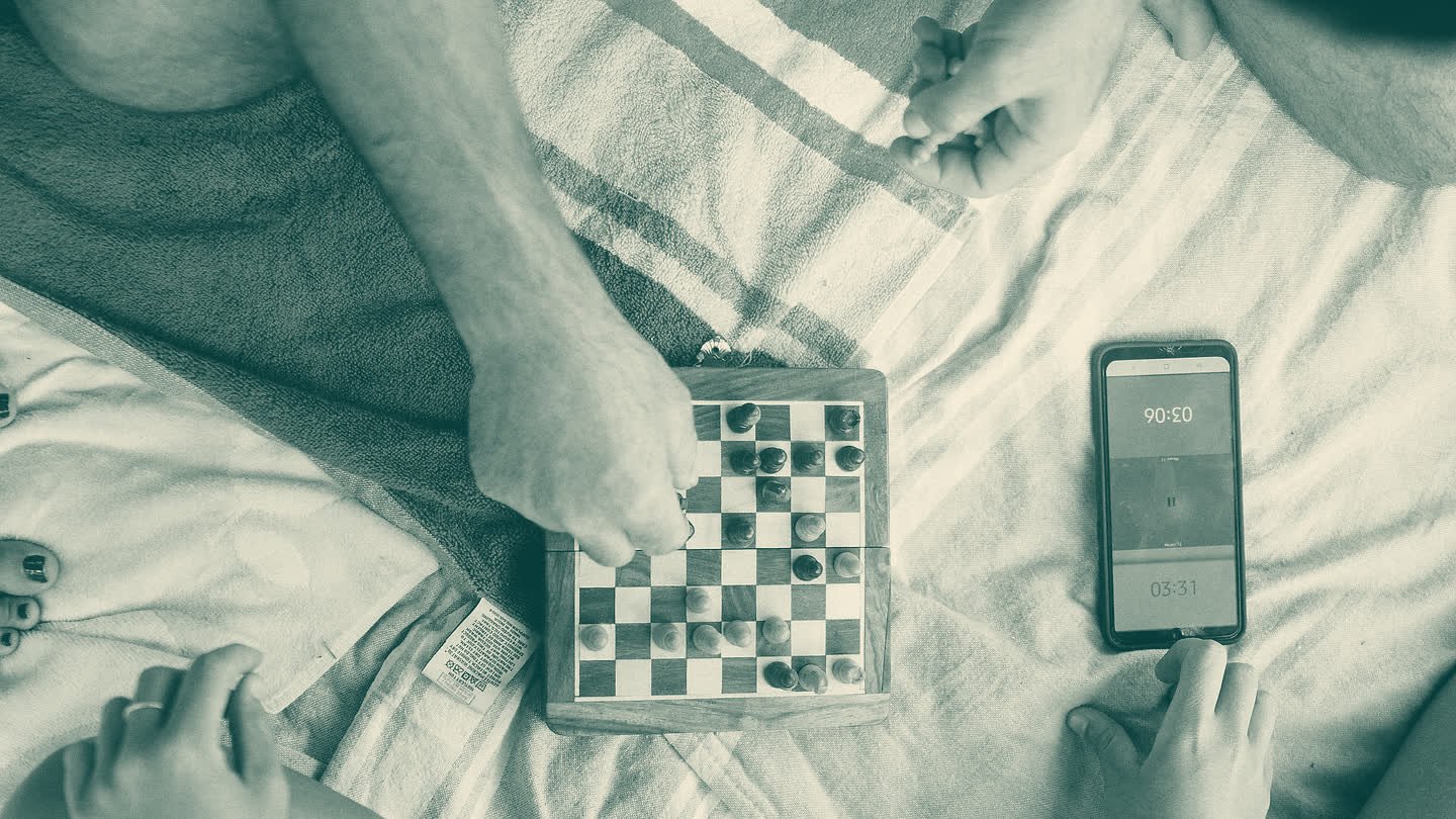 Seen from above: hands at a travel chessboard on a blanket, with a smartphone game clock beside it.