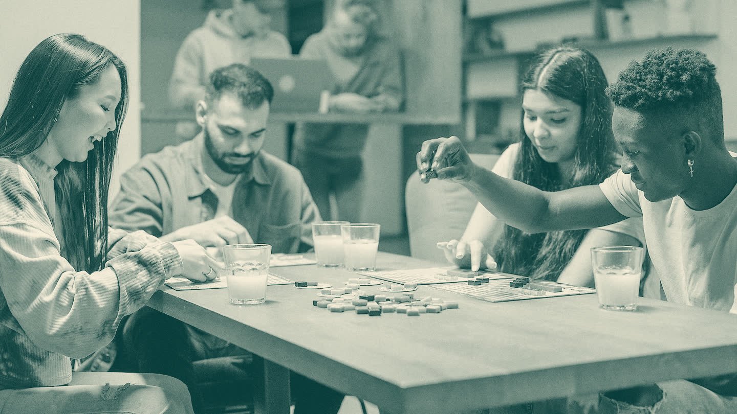 Four young adults play a tile-laying board game around a wooden table.